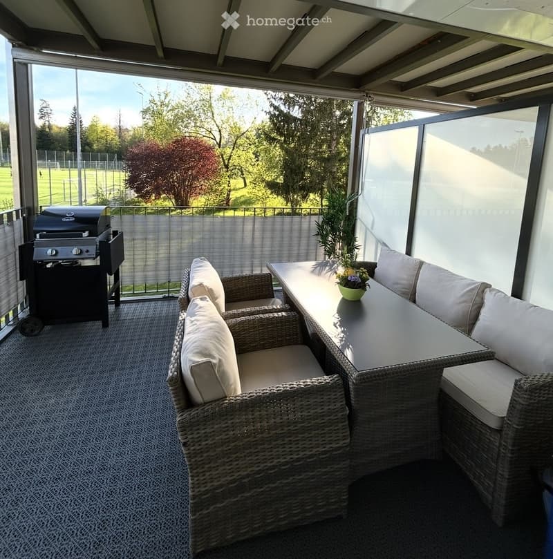 A covered patio with wicker furniture, a grill, and a small table with a flower pot, overlooking a green field.