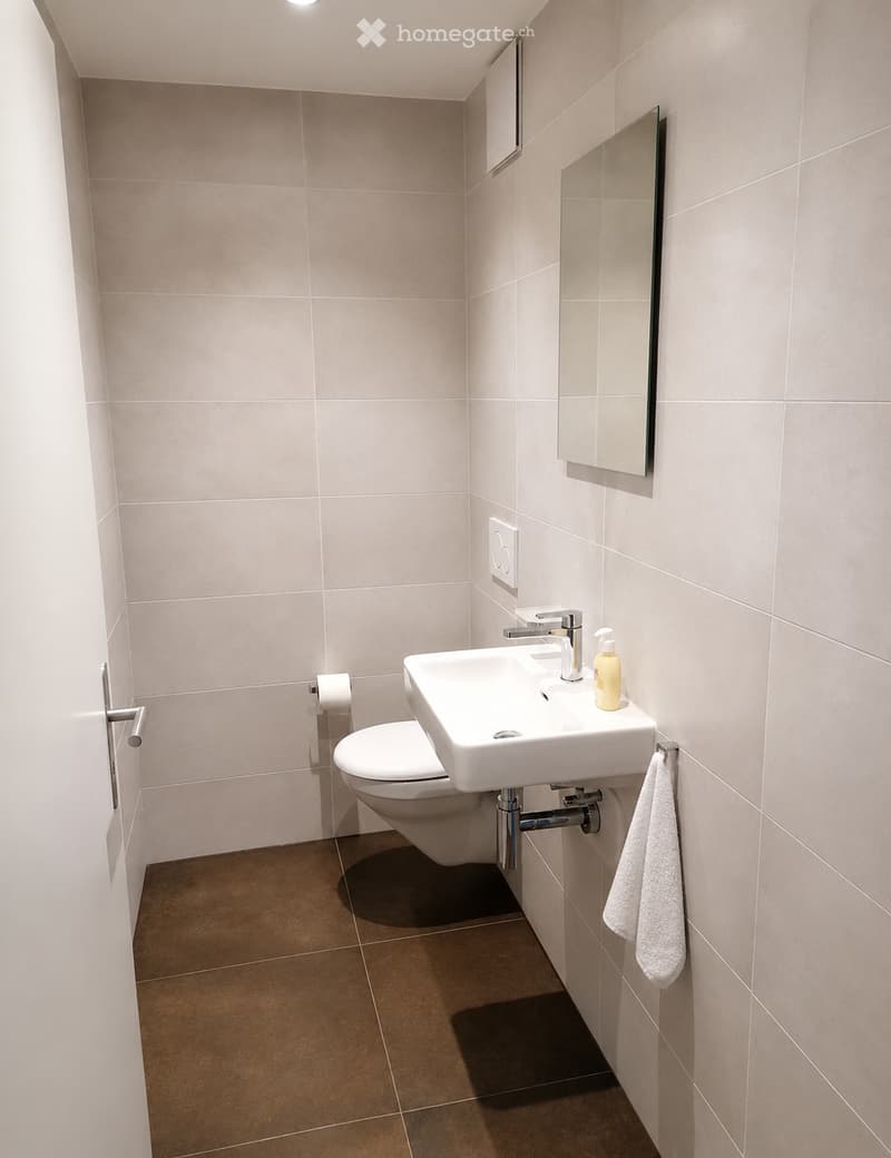 Modern bathroom with beige tiled walls, brown tiled floor, white toilet, white sink with chrome faucet, mirror above sink, and a white towel hanging on a rack.