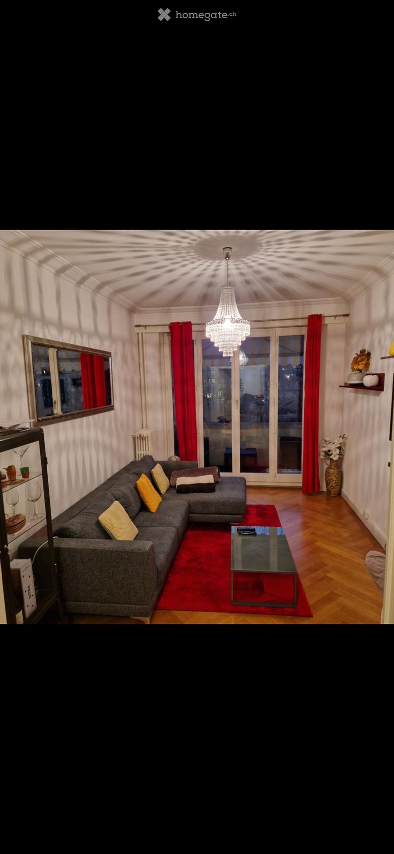 Modern living room with a grey sectional sofa, yellow throw pillows, a glass coffee table on a red rug, a chandelier, and large windows with red curtains.