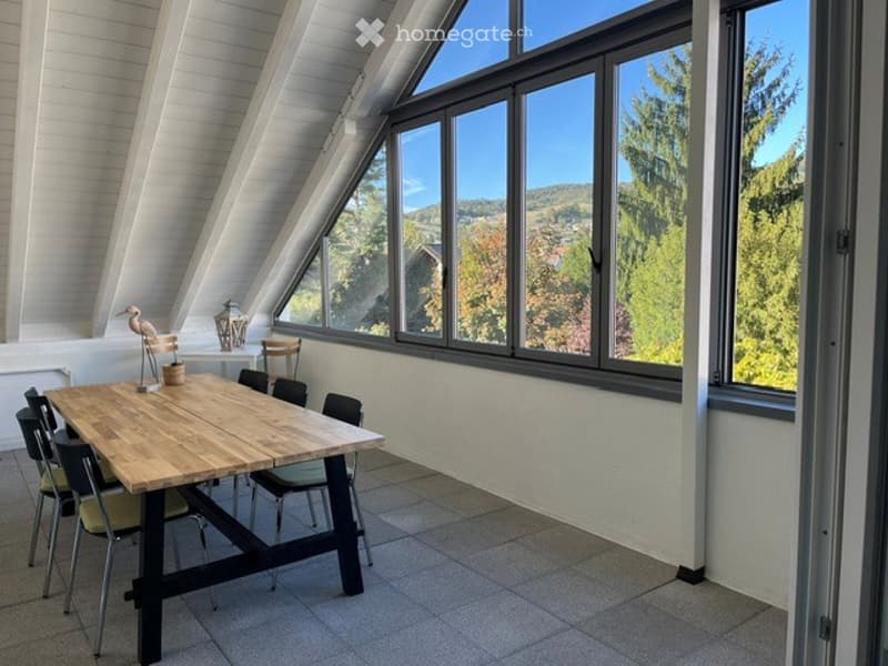Bright, airy room with large windows offering a scenic view of the landscape. The room features a wooden dining table with chairs and a decorative lamp on a side table.