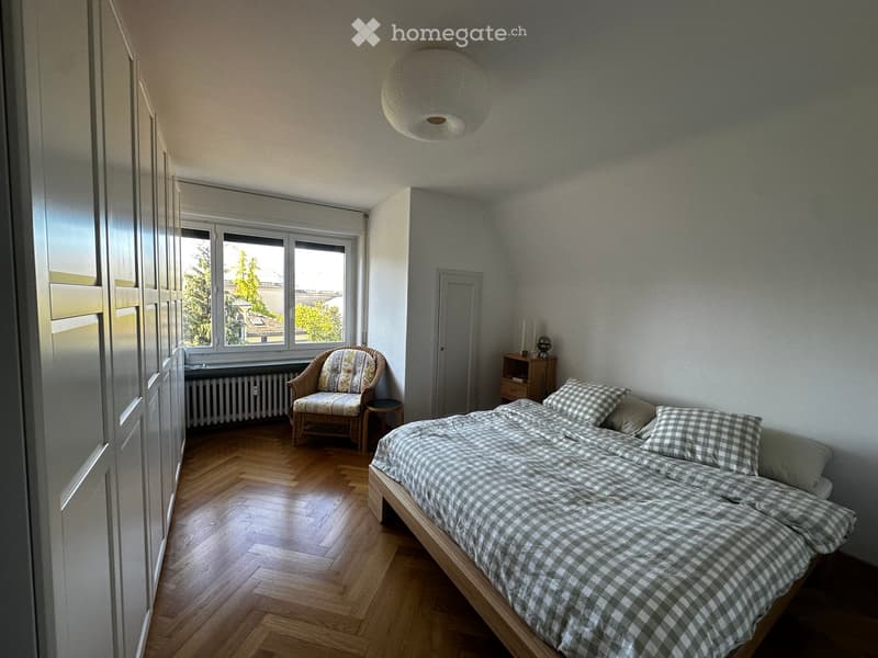 Cozy bedroom with wooden flooring, a double bed with checkered bedding, a wicker armchair, a small table, and a nightstand with a lamp.