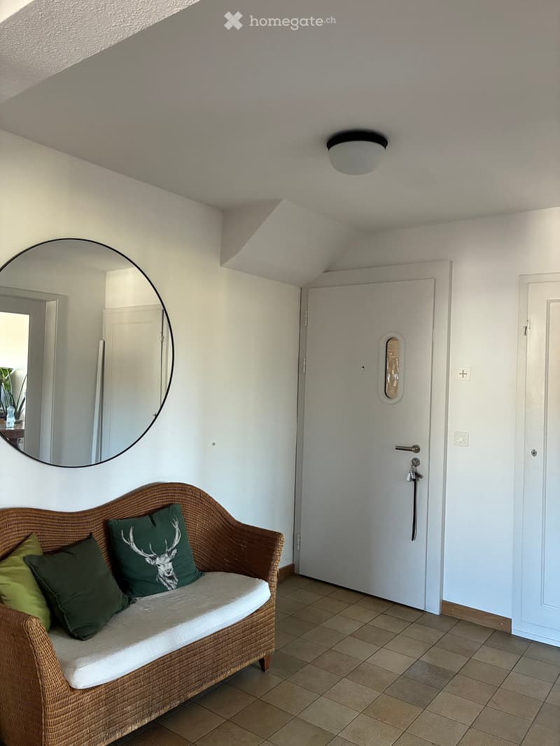 A small, cozy living space with a wicker sofa, green pillows, a large round mirror, and a white door with a decorative window.