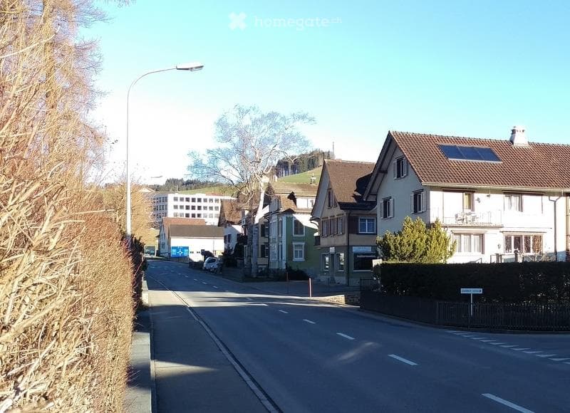 Residential street with multi-story houses, solar panels on roofs, parked cars, and trees lining the road.