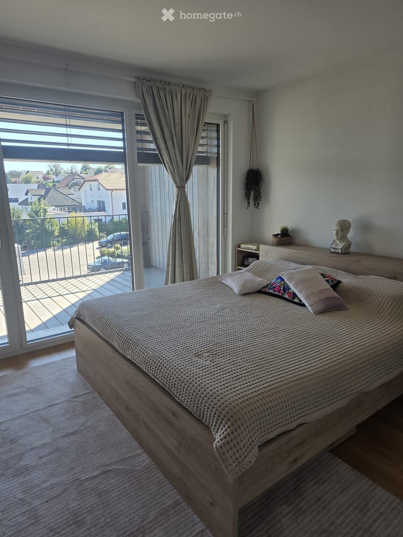 Modern bedroom with a wooden bed, decorative pillows, large window with balcony access, and a nightstand with a plant and sculpture.