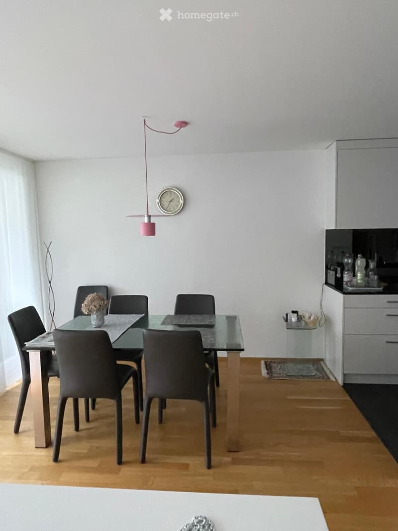 Modern dining area with a glass table, six black chairs, a vase with flowers, a clock on the wall, and a cabinet with drawers.
