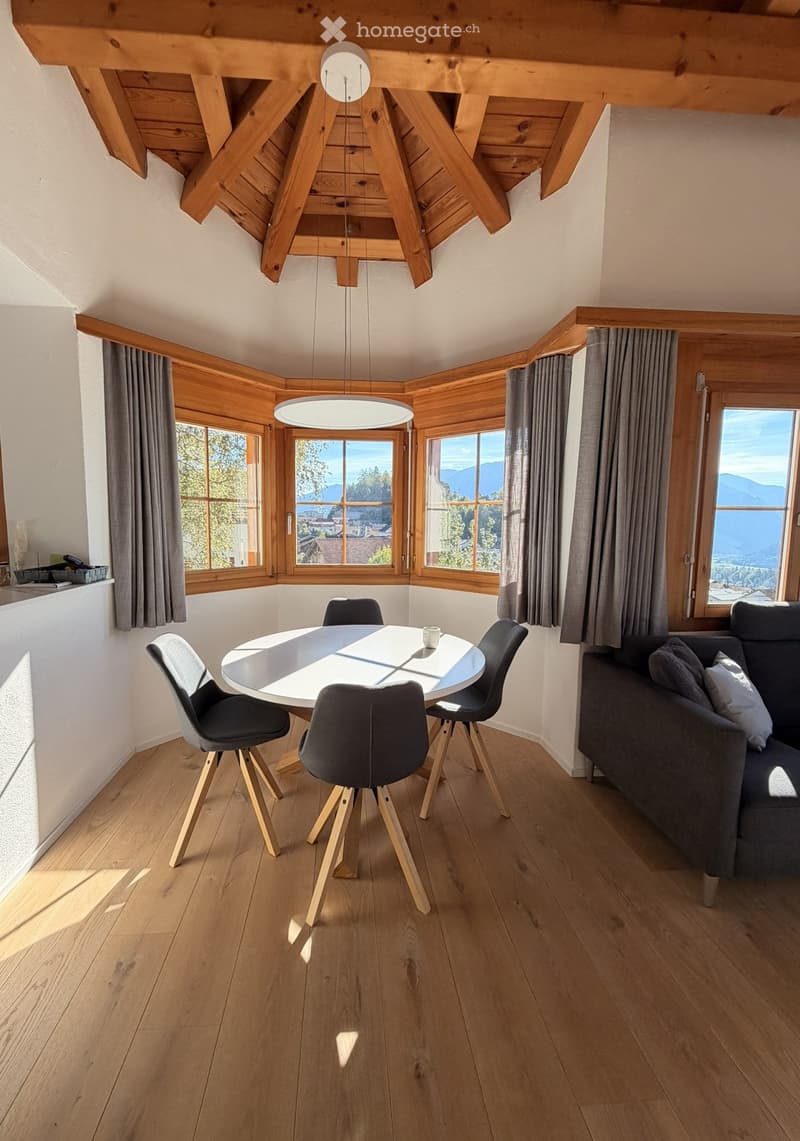 Modern living and dining area with wooden beams, large windows, a round dining table, and a grey sofa.