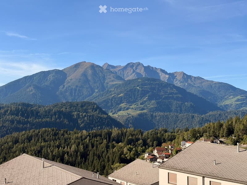 A panoramic view of a village nestled in a lush green valley surrounded by towering mountains under a clear blue sky.