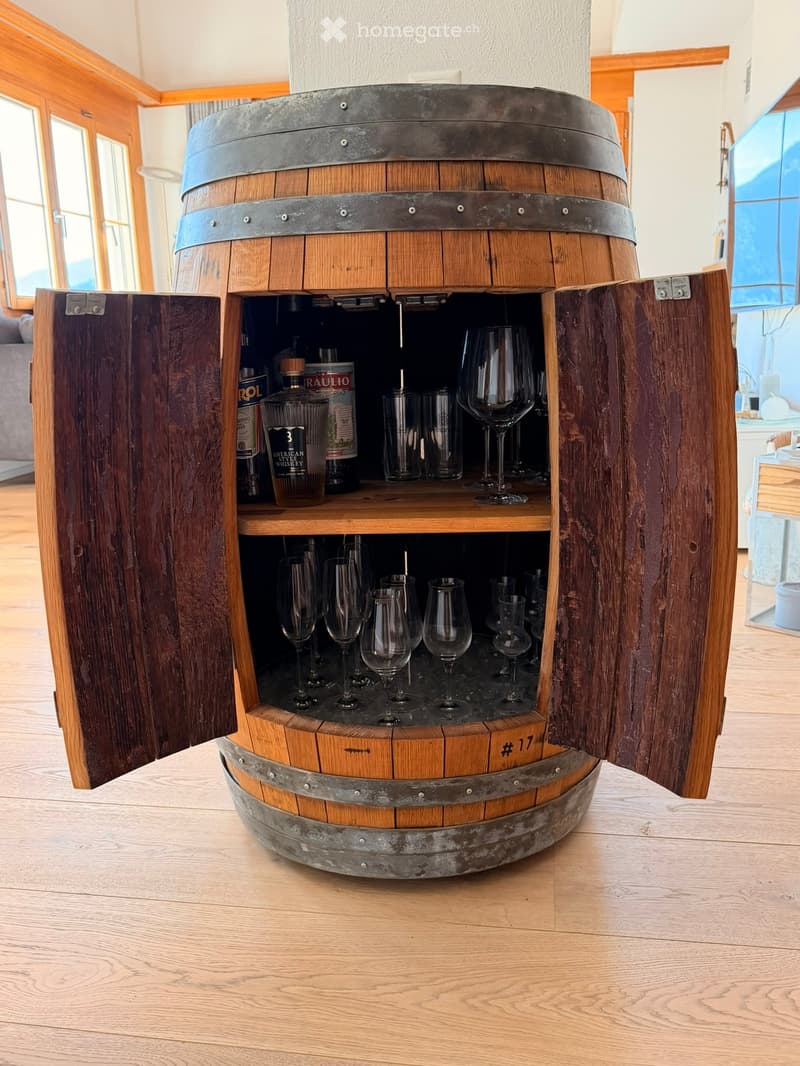 A wooden barrel-shaped bar cabinet with multiple shelves holding wine glasses and bottles, situated in a well-lit room with wooden flooring and large windows.