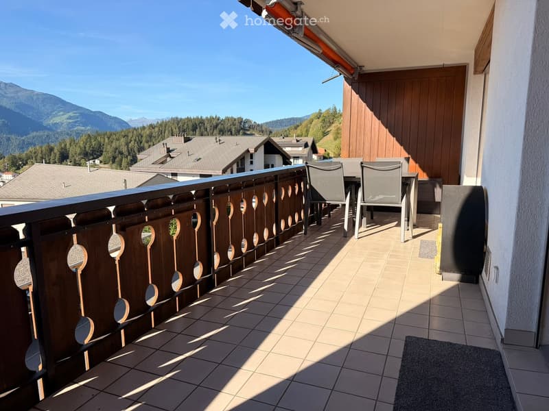 Balcony with tiled floor, wooden railing, and outdoor furniture including chairs and a table, overlooking a scenic mountain view.