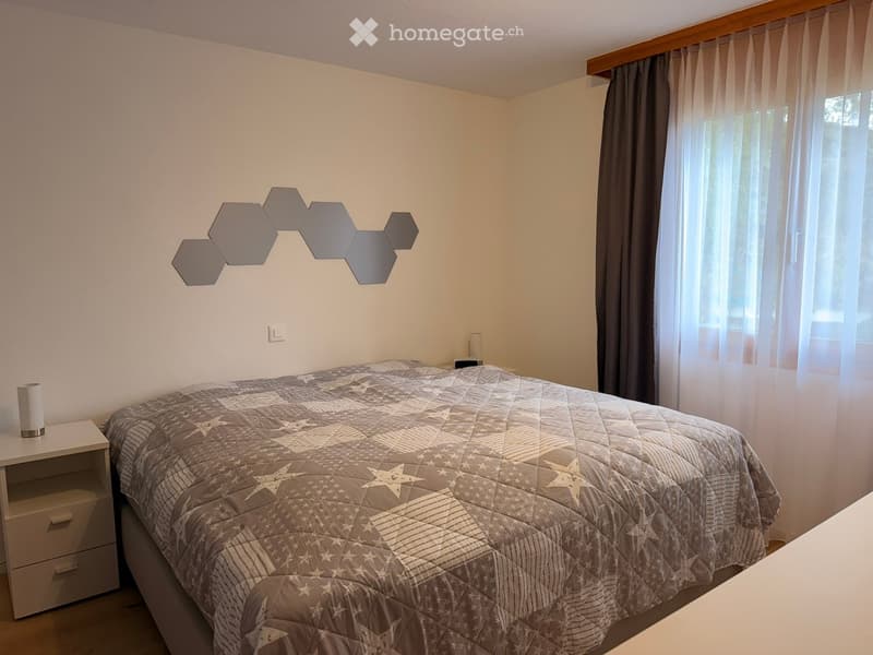 Bedroom with a double bed, nightstand, lamp, and decorative wall mirror. The bed has a quilted grey comforter with star patterns.