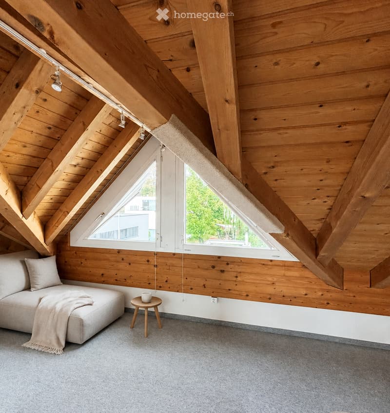 Modern attic room with wooden beams, exposed wooden ceiling, large skylight window, grey sofa, small wooden stool, and a cup on the stool.