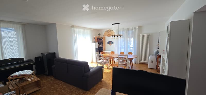 A spacious living and dining area with wooden flooring, a dark blue sofa, a dining table with chairs, a piano, and a fan on the wall.