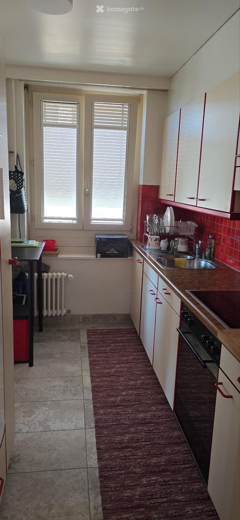 The kitchen features white cabinets with red trim, a wooden countertop, a sink with a faucet, and a dish rack. There is a radiator under the window, a small table with a red container, and a box on the windowsill.