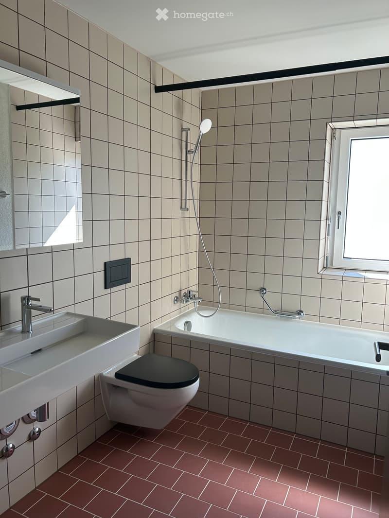 Modern bathroom with beige tiled walls, red brick floor, white sink, toilet, bathtub, and showerhead.