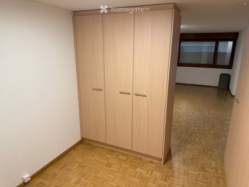 Empty room with wooden flooring, white walls, and a large built-in wardrobe with double doors. A window with blinds is visible in the adjacent room.