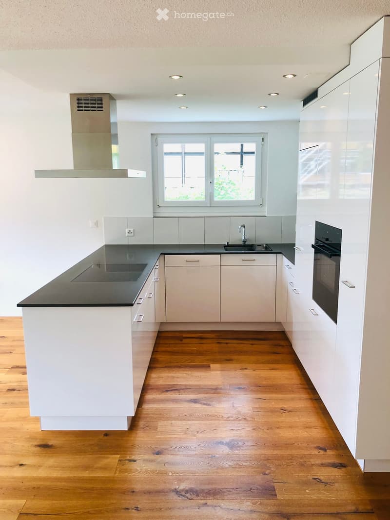 Modern kitchen with white cabinets, black countertops, wooden flooring, and a large window.