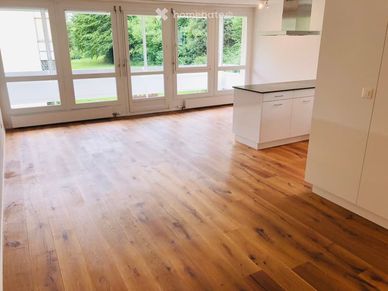 Empty room with light wooden flooring, white walls, large windows, and a small white kitchen unit in the corner.