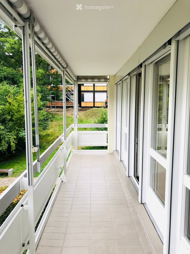 Balcony with white tiled floor, white railings, large windows, and a view of greenery and a building in the background.