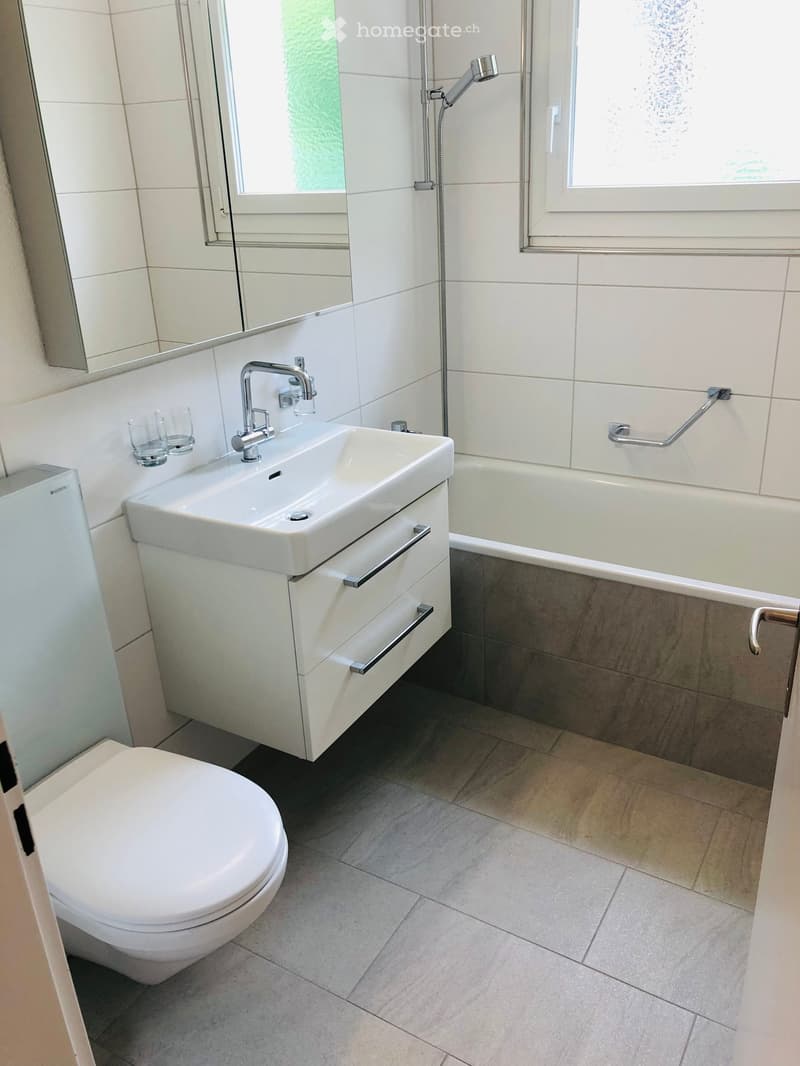 Modern bathroom with white tiles, a toilet, a sink with a modern faucet, a bathtub, and a mirror above the sink. Two glasses are placed on the sink counter.
