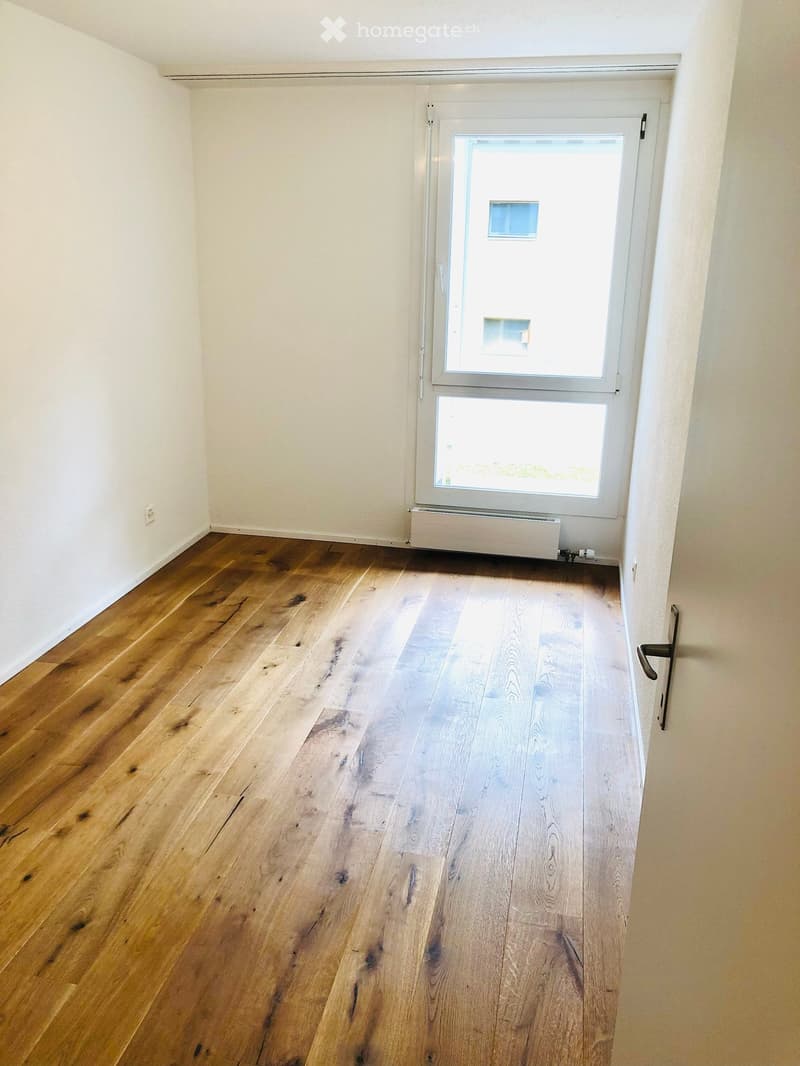 Empty room with wooden flooring and white walls, featuring a large window with white frame and a radiator below it.