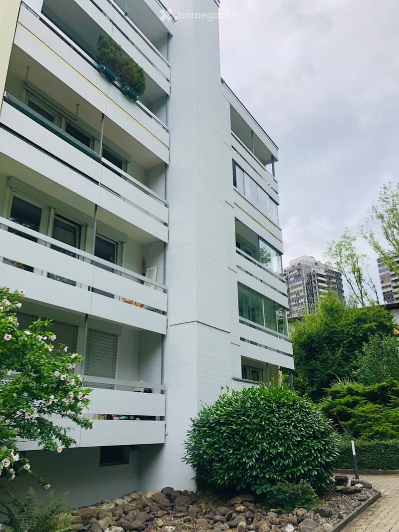 Modern white apartment building with multiple balconies and large windows, surrounded by greenery and rocks at the base.
