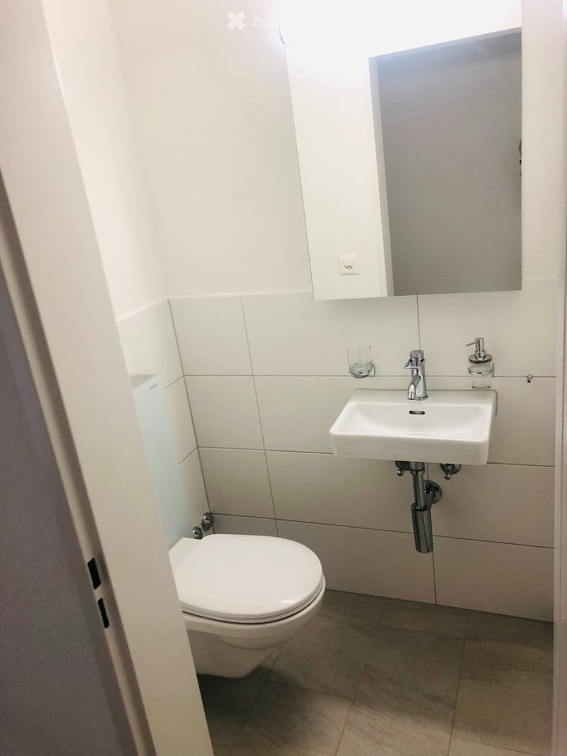 Modern bathroom with white tiles, a toilet, a sink with a chrome faucet, a mirror, and a soap dispenser.