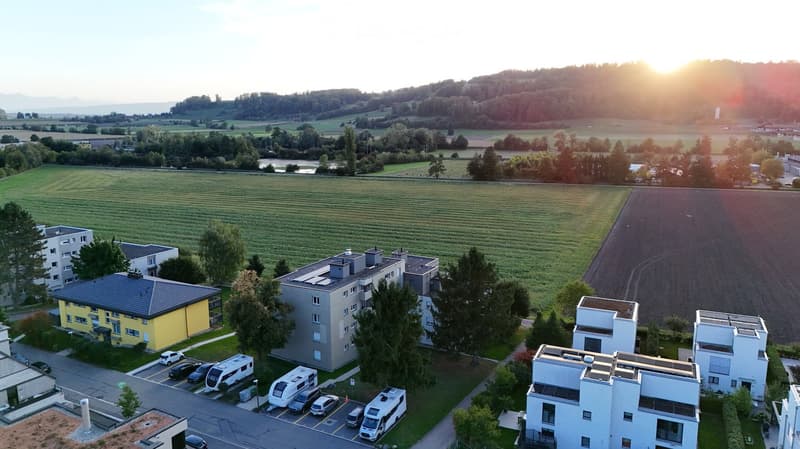 Aerial view of a residential complex with multiple buildings, surrounded by green fields and trees. Several cars and RVs are parked in the lot.