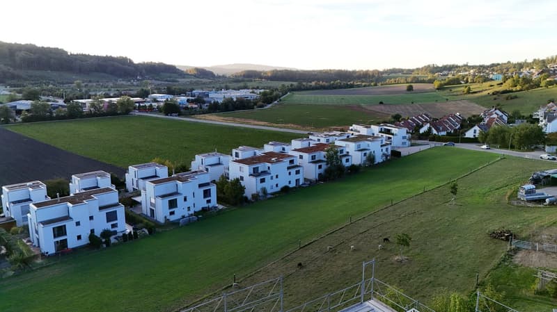 Aerial view of a residential area with modern white houses, surrounded by green fields and a rural landscape.