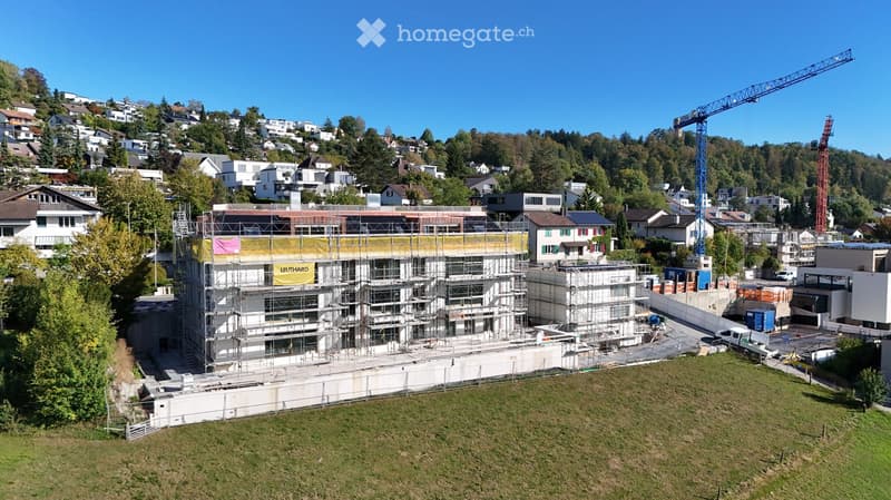 A multi-story residential building under construction, surrounded by scaffolding and cranes, with neighboring houses and greenery in the background.