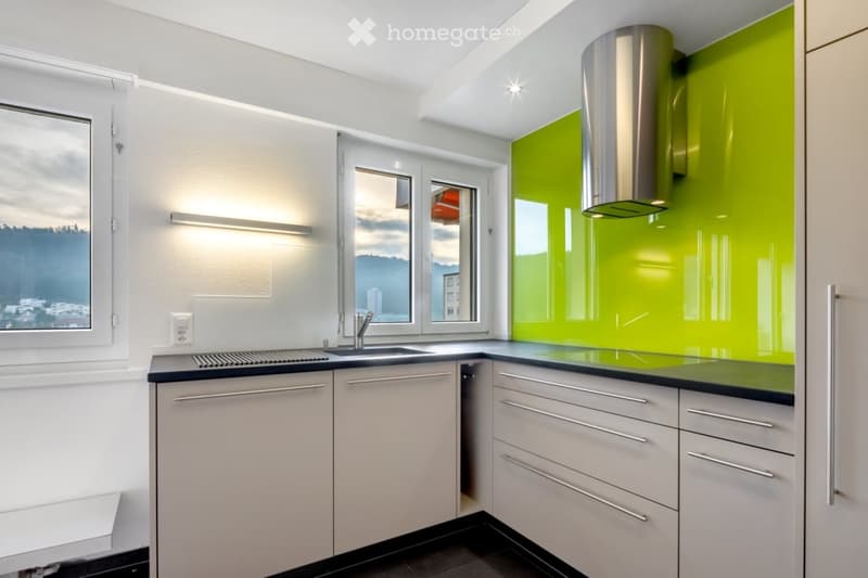 Modern kitchen with white cabinets, black countertops, stainless steel range hood, and a green accent wall. Features a large window with a scenic view.