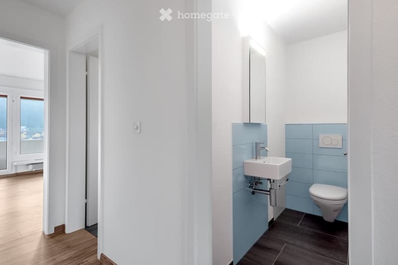 Modern bathroom with white walls, light blue tiles, a white sink, a toilet, a mirror, and a cabinet. The floor is wooden, and there is a view through an open doorway.