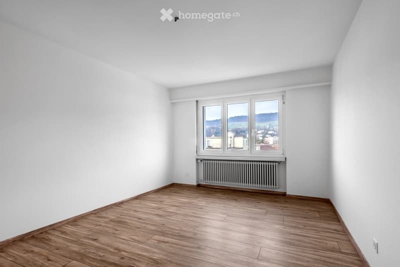 Completely empty room with white walls, wooden floor, and a large window with a view of the landscape.