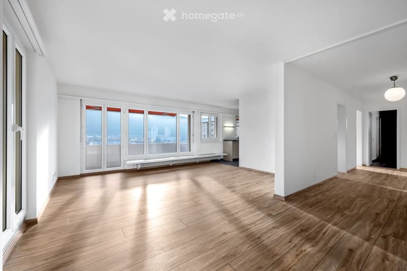 Bright, unfurnished room with wooden flooring, large windows offering a city view, and white walls.