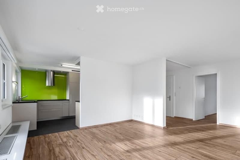Bright, unfurnished living space with wooden flooring, white walls, and a small kitchen area featuring a green accent wall, sink, and cabinets.