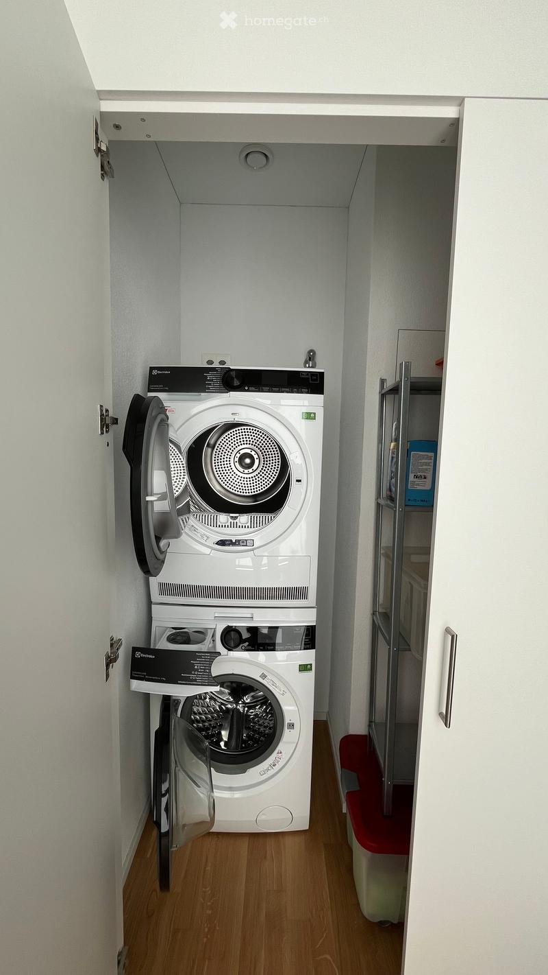 Compact bathroom with a stacked washing machine and dryer unit, white walls, wooden floor, and a small red waste bin.