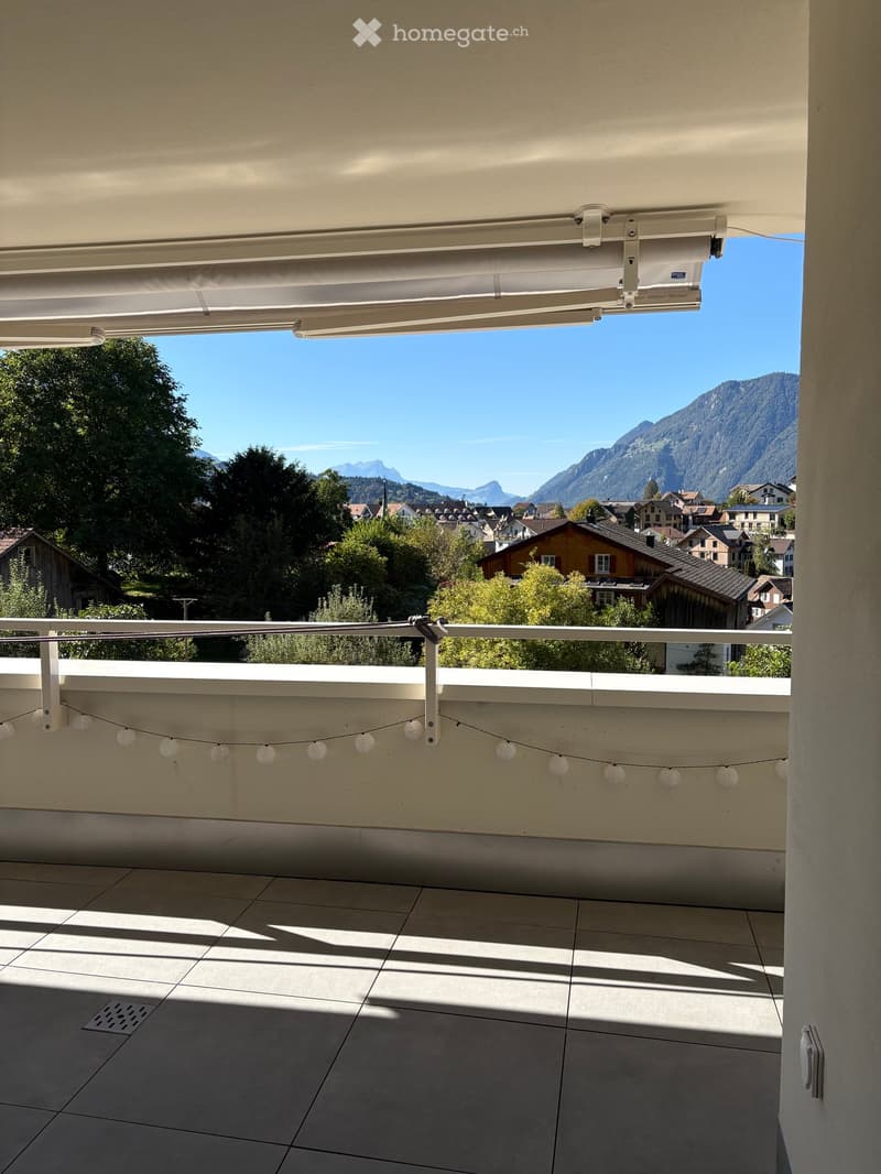 Balcony with tiled floor, white railing, string lights, and a retractable awning, overlooking a town and mountain landscape.
