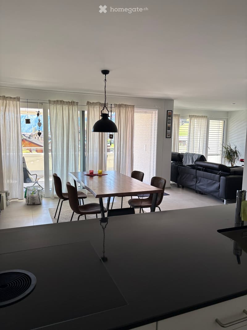 Open-plan living and dining area with a modern kitchen counter, a dining table with chairs, and a black leather sofa. Large windows with curtains and a view of the outdoor area.