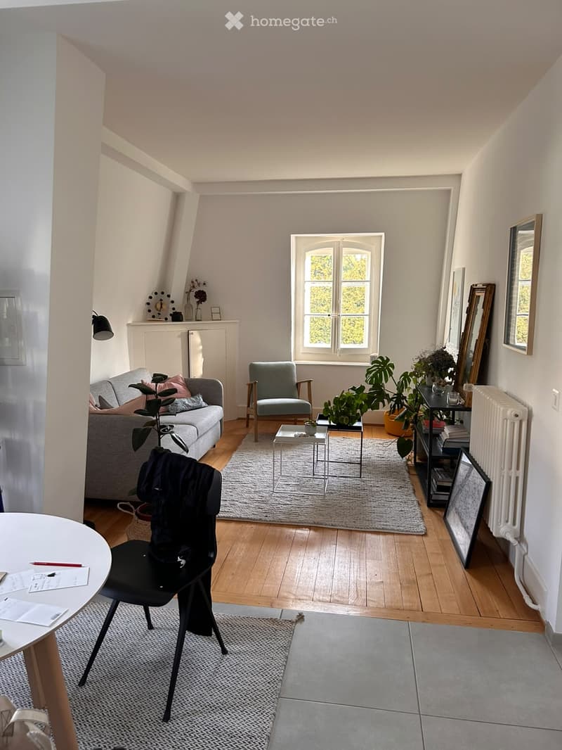 Bright living room with wooden flooring, large window, grey sofa, armchair, coffee table, and several plants. Includes a small dining area with a round table and chair.
