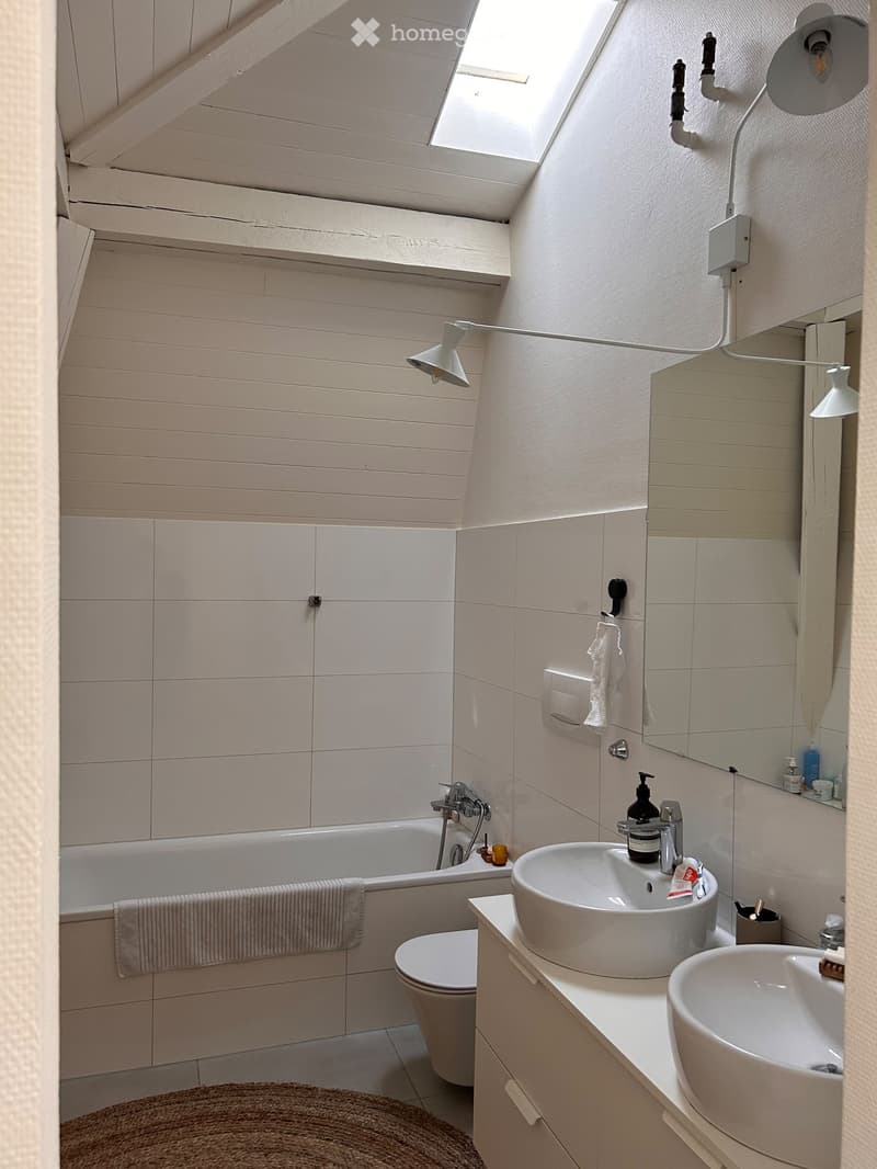 Modern bathroom with a bathtub, toilet, double sink, mirror, and wall-mounted light fixtures. The space features white tiles and a skylight window.