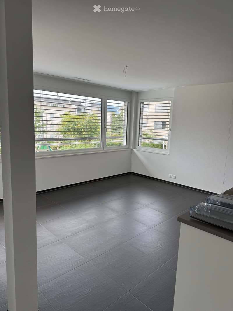 Empty room with dark tiled floor, white walls, large windows with blinds, and a small black countertop with a glass object on it.