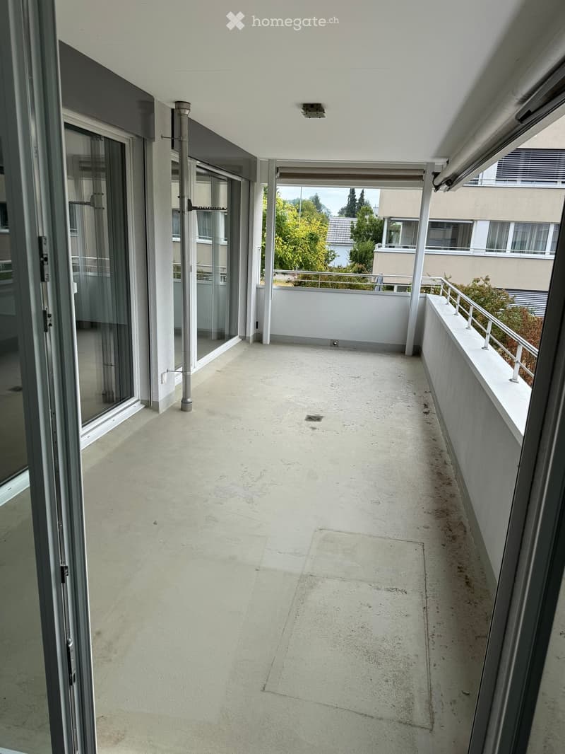 Balcony with concrete floor, white walls, metal railing, and sliding glass doors leading to the interior.