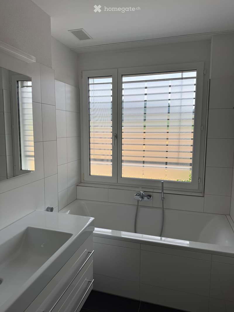 Modern bathroom with a white bathtub, sink, and faucet, featuring large windows with blinds and white tiled walls.