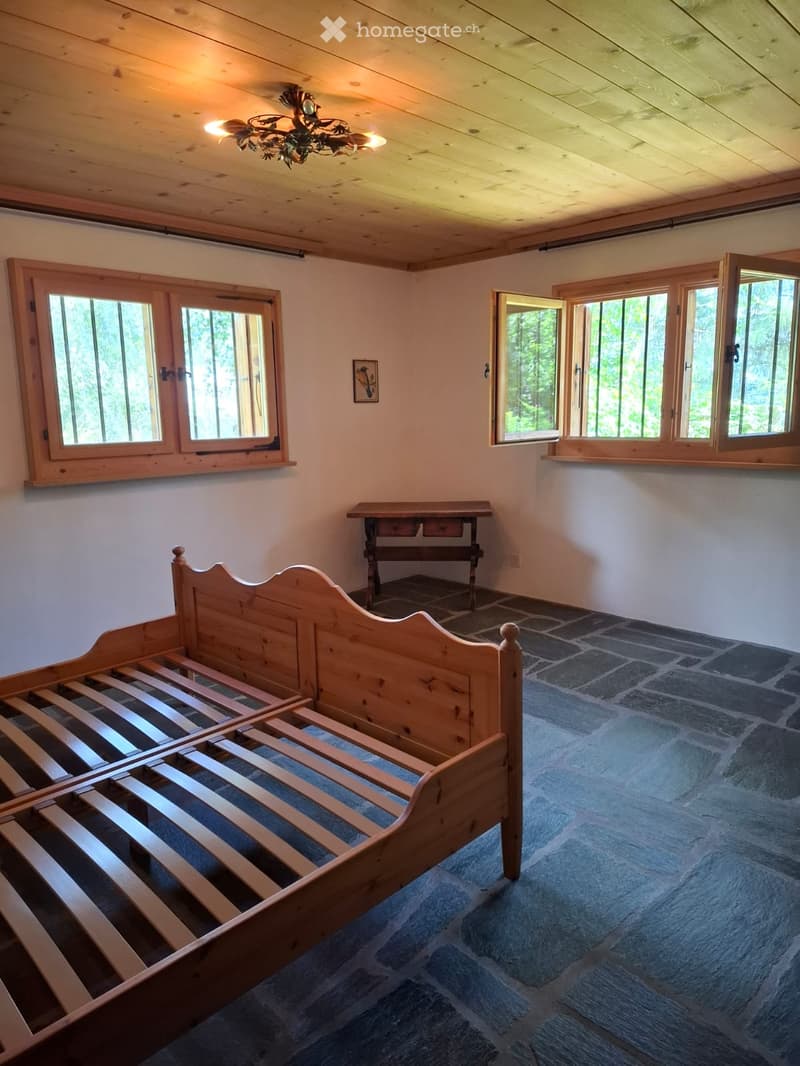 Cozy bedroom with wooden bed, small table, chandelier, and large windows overlooking a forested area.