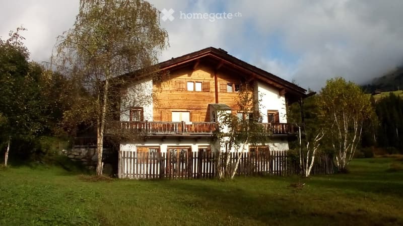 A two-story wooden house with white walls, multiple windows, and balconies, surrounded by a wooden fence and trees in a grassy area.