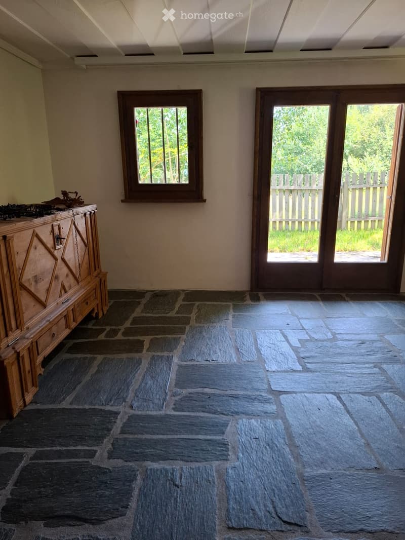 An unfurnished room with a stone tile floor, a large window, and a wooden cabinet with a decorative sculpture on top. The room has a high, slanted ceiling and a sliding glass door leading to an outdoor area.