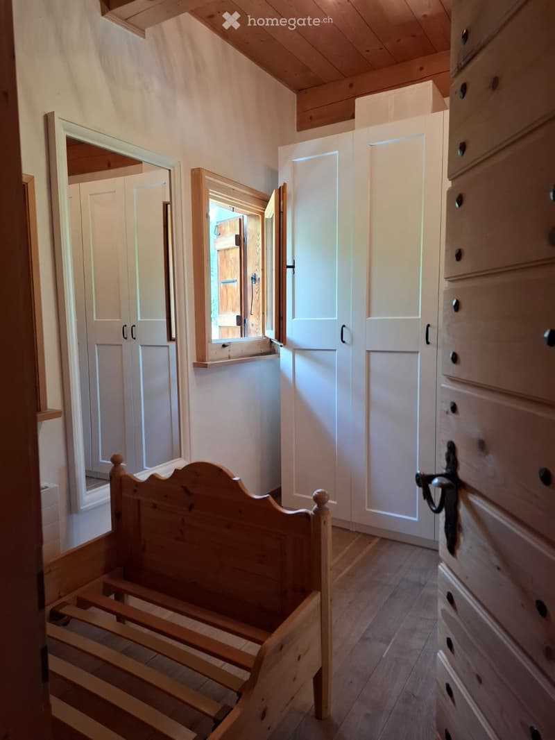 Empty room with wooden floor, exposed wooden beams on the ceiling, built-in white cabinets, and a partially assembled wooden bed frame.