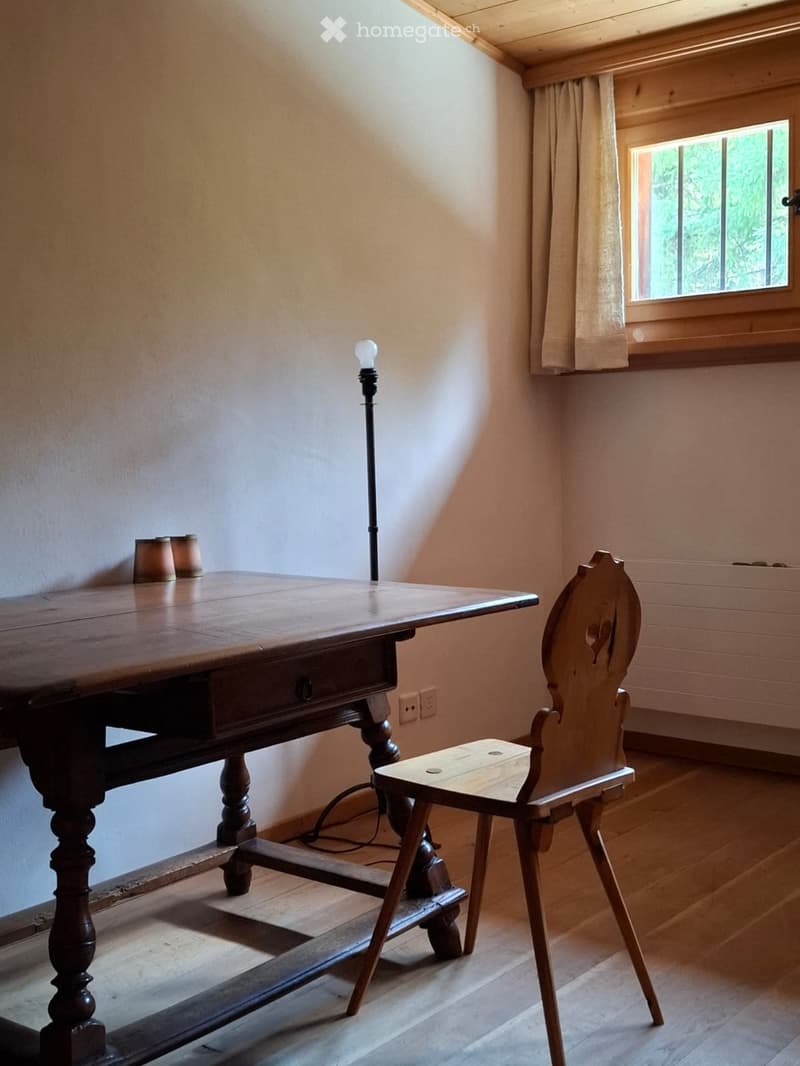 A small, cozy room with wooden flooring, a wooden table, a single chair, and a floor lamp near the window.