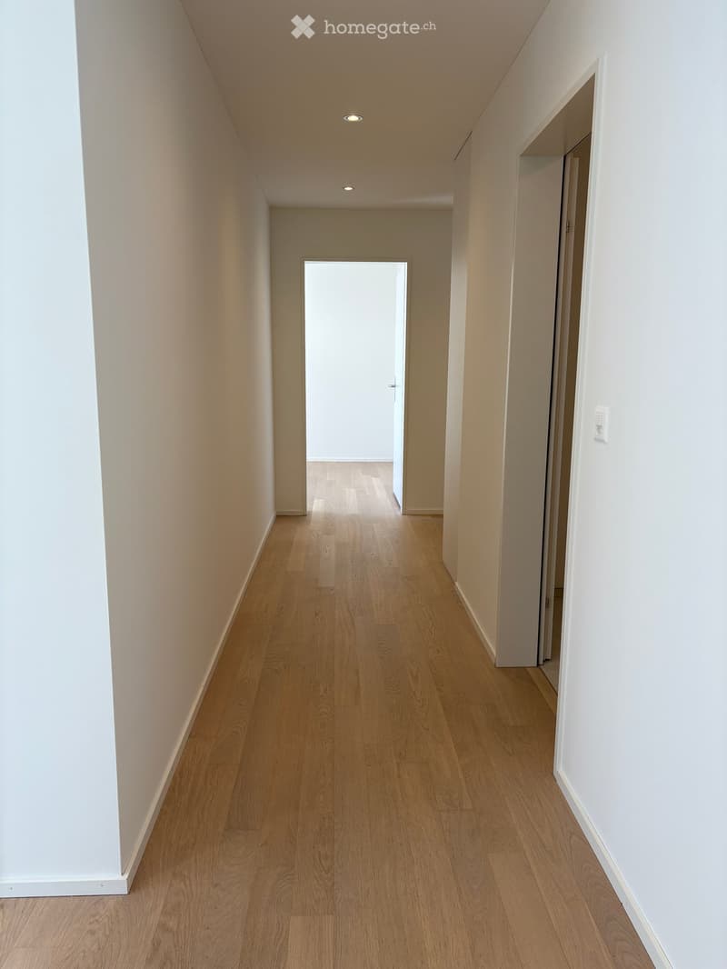Empty hallway with light wooden flooring and white walls, featuring recessed ceiling lights and an open doorway at the end.
