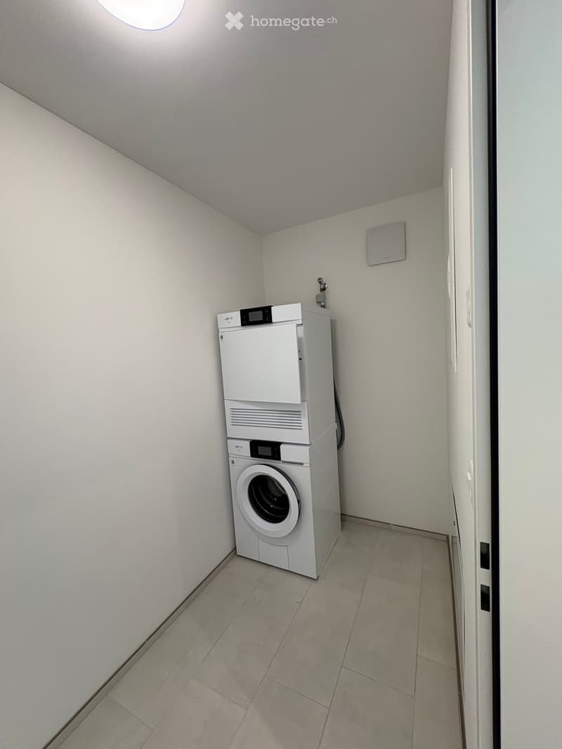 A small, empty room with light-colored tiled flooring and white walls. A white washing machine and dryer are stacked against one wall, and a door is visible on the right side.