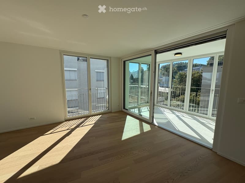 Empty room with wooden flooring and large sliding glass doors leading to a balcony. Sunlight streams in, casting shadows on the floor.
